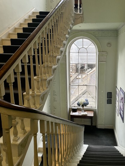 A historic staircase with white banisters and a dark carpet runner leading up to a large arched window. A small table with flowers and books sits in front of the window, and posters or artwork are displayed on the wall to the right.