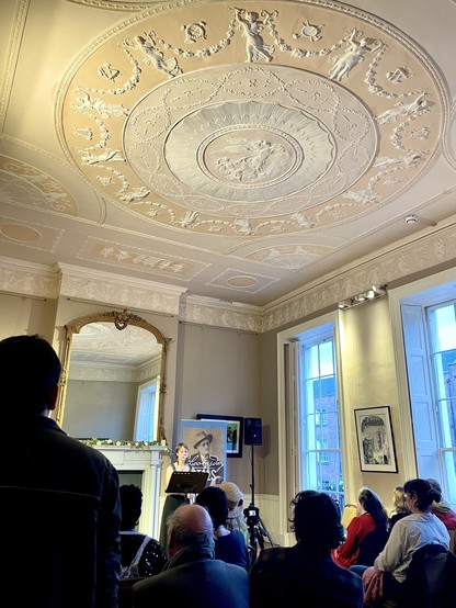 A speaker presenting in a beautifully decorated room with an ornate plaster ceiling and an audience seated and listening attentively.