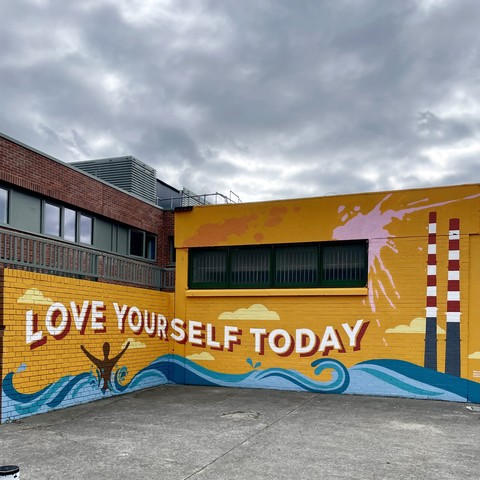 Colorful mural with the words "LOVE YOURSELF TODAY" on a yellow brick wall, featuring a person raising their arms amidst waves and Dublin’s two iconic Pigeon House  industrial chimneys in the background.