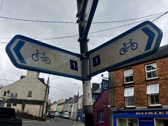 Two Euro Velo 1 signs at a junction, ninety degrees to each other. Behind it you see the main road that leads up the hill to the 200 meters of cycle lane shown in the first photo.