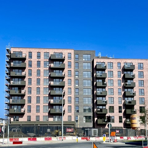 A modern apartment building featuring multiple balconies and large windows. The structure has a mix of brick and dark-colored panels, with construction scaffolding on the left side. The sky is clear and blue. Traffic signals and signs are visible in the foreground.