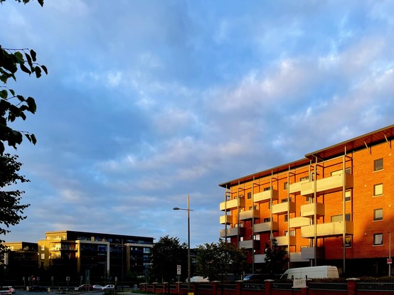 A street view featuring modern apartment buildings bathed in warm sunlight against a partly cloudy sky, with trees and vehicles visible in the foreground.