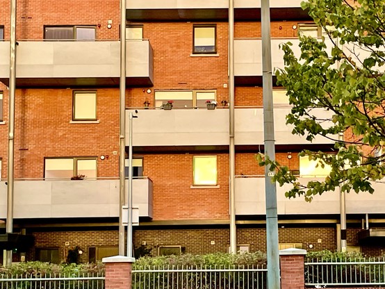 A multi-story residential building with brick and concrete facades. Several balconies are visible, featuring potted plants. A streetlight and some greenery are in the foreground.