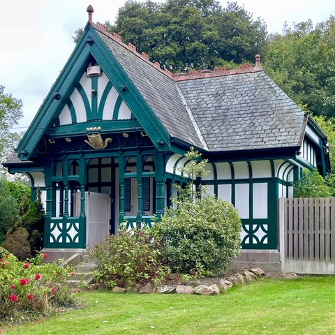 A small, ornamental building featuring intricate green and white wooden structures, a gabled roof with slate tiles, and decorative detailing. Surrounded by neatly kept grass and foliage, the building is set against a backdrop of trees.