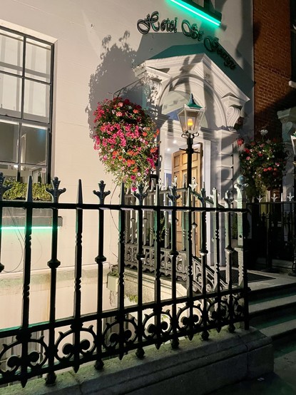 A nighttime view of the entrance to Hotel St. George, featuring a decorative facade, hanging flower baskets, and a lamppost. The building is illuminated by soft lighting, highlighting the architectural details. A wrought iron fence runs along the front.