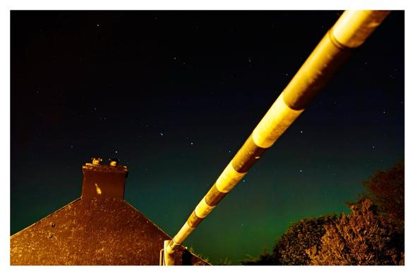 Last of the green glow in the night sky over a house, trees and a black and yellow entrance barrier cuts across the image.