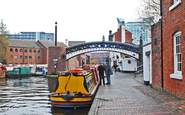 A view of a canal basin with several narrowboats and red-brick buildings in the background. There is a small pretty bridge. It is painted white with black railings along the side. It appears to start halfway up the buildings on the right of the picture, cross over the canal footpath and land on a brick structure in the middle of the water. A bright yellow houseboat is squeezing through underneath it.