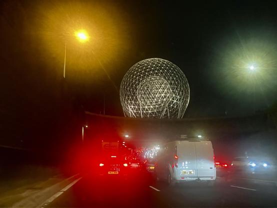 Arriving in to Belfast on the motorway. A wire sphere towers above the road Red, yellow, blueish and greenish lights of the street and traffic against the night sky