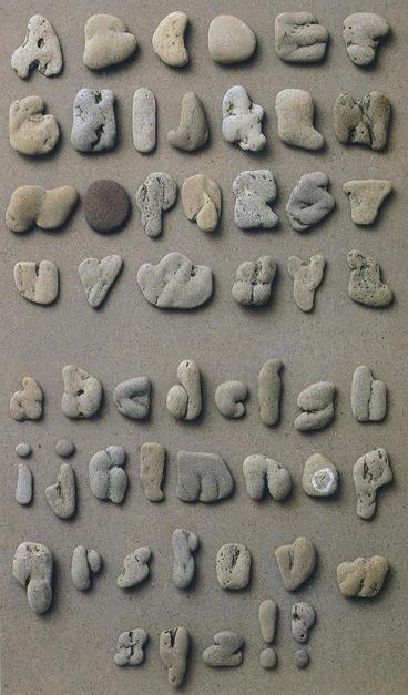 A typeface set that consists of uppercase then lowercase letters, followed by an exclamation mark and question mark, placed on a light-grey surface. Each one is a pebble resembling the letter or mark in question. Some of the resemblances are impressive, and the overall effect is marvellous. The pebbles are mostly shades of grey, many tinged yellow or brown, and their textures give them great variety of character and appearance.