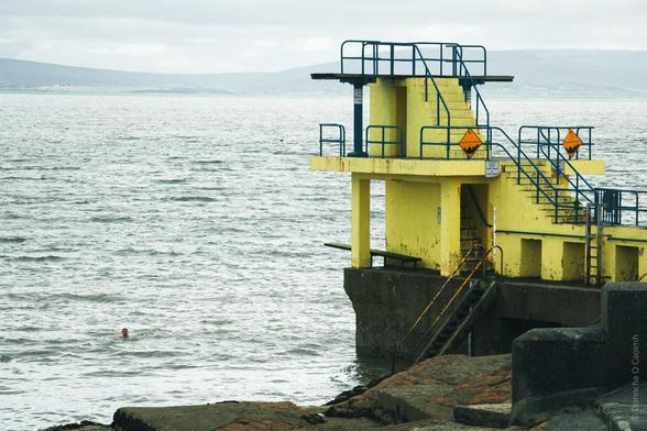 A yellow diving platform or swimming structure extends into choppy grey waters on an overcast day. The platform features blue railings, safety warning signs, and multiple levels accessible by metal stairs. A lone swimmer can be seen in the water near the platform. The scene is set against a backdrop of distant hills or cliffs shrouded in mist, and rocky shoreline is visible in the foreground.