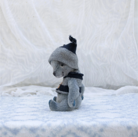 Beautiful handmade teddy bear. It's silver-gray with delicate airbrushed shading that makes him look almost alive. He's wearing a gray and black jacket and a hat, and smiling. He's photographed on a fluffy white background adding to the gentle winter feeling 