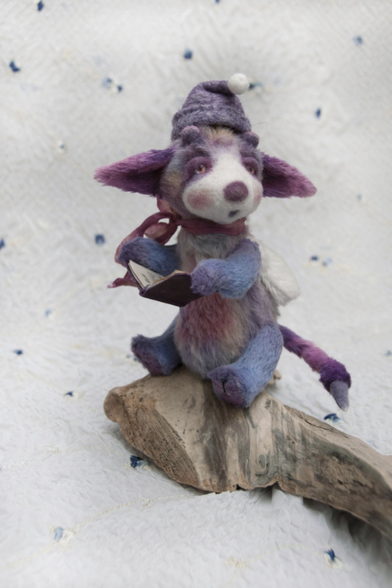 Another handmade custom teddy but it looks not much like a bear, but a fantastical creature. It's purple and blue, has a tail, and pointy ears to the side. His face is in a singing expression and he's holding a miniature lyrics book. He's also wearing a winter hat and is photographed sitting on a stone on a soft white background, giving illusion of snow. 