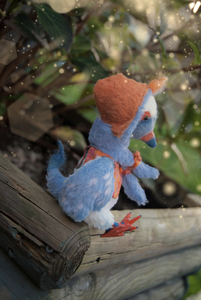 Another fantastical creature, in a shape of a bird. It's predominantly blue but with orange elements, such as its duck like legs, beak, and shading around the eyes. He's wearing an orange waistcoat and an orange hat and sitting on a wooden frame with blurred greenery behind it. 
