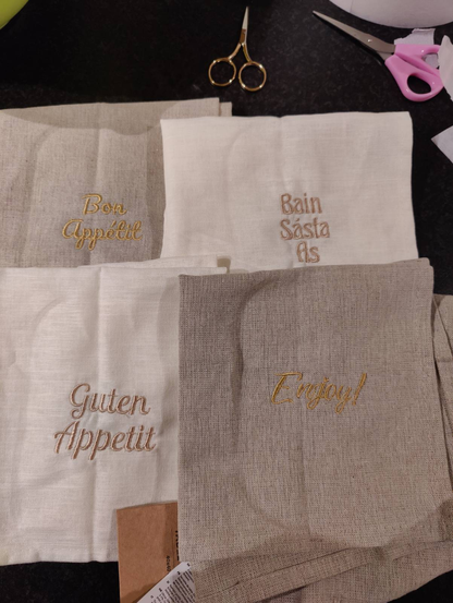 Four napkins lying on a black counter with scissors nearby. Two are stone with gold writing and two are cream with rose gold writing. Each of them has a different message, namely: "Bon Appétit", "Bain Sásta As", "Enjoy!", and "Guten Appetit".