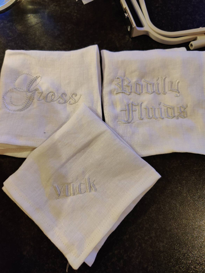 Three white linen hankies, folded, and laid out on a black counter. One has "Gross" embroidered in a curly font. Another has "yuck" in an art deco ish font. The third has "Bodily Fluids" in a medieval manuscript font. The corner of an embroidery hoop is visible in the corner.