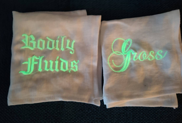 A slightly blurry photo of two of the aforementioned hankies in a darkened environment. In green glow in the dark font they read Bodily Fluids and Gross.