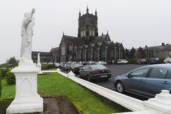 Gothic church exterior with large car park and white religious statues in the foreground at Mount Melleray Abbey, showing dark stone architecture and central tower under overcast sky.
