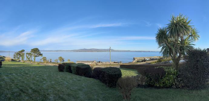 Mid morning panorama of Dungarvan bay viewed from Helvick looking north across a dead calm sea to the Comeragh plateau. 

The sky is a bright blue with streaks of very wispy clouds crossing from west to east 

A frosty lawn with some bushes is visible in the shaded foreground There are fir trees in the middle distance on the left of the picture down by the sea and a cordalyne Australensis on the right just  a raking up into the low morning sunlight. 