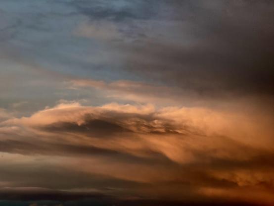 Dramatically lit dark orange rain cloud with lots of flowing details and ripples against a very dark blue sky with the tendrils of a looming black rain cloud extending in from the right of the picture. 