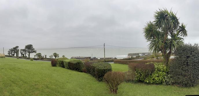 Mid morning panorama of Dungarvan bay viewed from Helvick looking north across a wild stormy sea. 

The grey sky and rain almost completely obscure the far side of the bay and the Comeragh Plateau beyond. The water is extremely choppy with lots of white capped waves and long streaks of foam from long breakers rolling in from the east. 

A green lawn with some bushes is visible in the shaded foreground There are fir trees in the middle distance on the left of the picture down by the sea and a cordalyne Australensis on the right that is being tossed about in the high wind. 