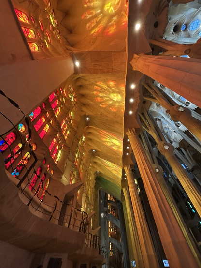 Photo of the interior of La Sagrada Familia church. The widows are mostly red yellow and orange stained glass. The bright sun is shining through the windows making a reflection on the ceiling of the church. Opposite the windows are tall columns and pillars that look like trees in a forest. 