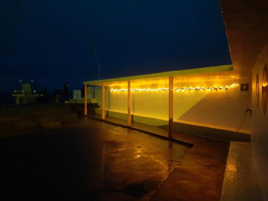 Photo of a concrete bathing shelter painted yellow with red columns. A line of fairy lights under the canopy casts a soft light onto the wall and the wet concrete floor. The diving tower can be seen dimly in the distance on the left.