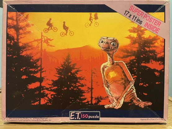 A vintage puzzle box featuring E.T. the Extra-Terrestrial. The artwork showcases the alien character alongside children riding bicycles against a vibrant sunset backdrop with silhouettes of trees. The box indicates it's a 150-piece puzzle and includes a super poster inside.