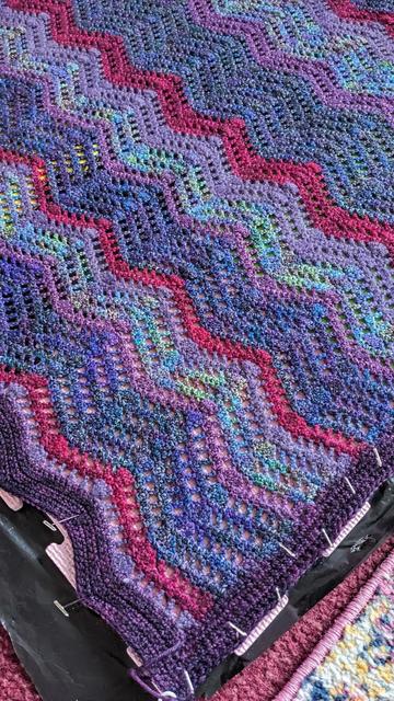 Close up on a blanket which has been pinned out for blocking. The blanket is crocheted in a lacy ripple stitch, in rows of different colours of purples and plums.  A thin ruby coloured row provides contrast every now and then.