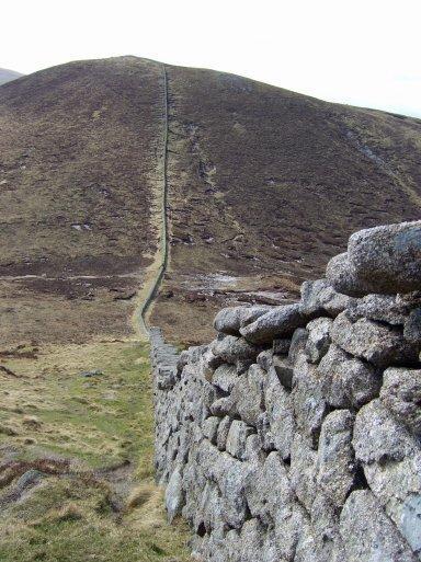 A grey granite dry stone wall stretches across a scrubby heather covered plateau, climbing up the slope of the mountain in the distance.