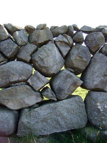 A straight on look at the intricate construction of a granite dry stone wall, showing the seemingly random boulders that make up the main body of the wall. The top level of the wall is capped with much smaller stones. The grass of the field behind the wall can be seen in the gaps between the stones.