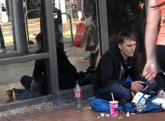 street scene of a person in a black top sitting in front of soft-drink bottles and cups and food packets, reflected in the glass windows behind; on the far right a pedestrian in orange top passes by