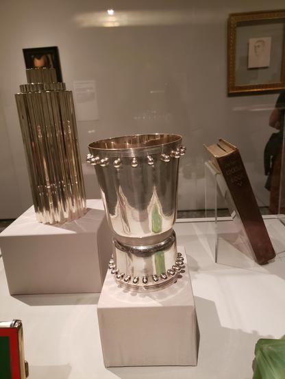 Small display of objects from Lempicka (art deco painter) exhibit. A silver thin tall rippled object and a book and in the center a gorgeous Jean Puiforcat silver goblet. It has a lower cylinder base with tiny silver balls around the edge. The upper part is slightly larger around and has tiny silver balls around the top. The front right has green reflections of a glass object (not in picture).