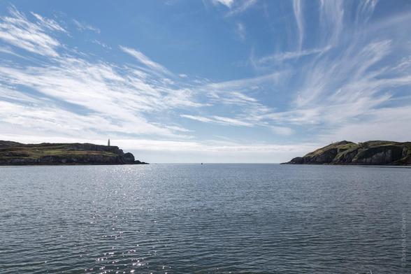 text
A serene view of the sea channel leading to Sherkin Island, Cork, Ireland, with calm waters reflecting sunlight and rugged cliffs framing the scene under a bright blue sky scattered with wispy clouds.