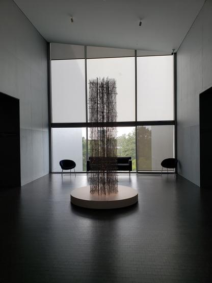 A huge window with 2 black chairs and a black sofa in front. There is a round base about 10 feet in front of it, with a tall (15 feet?) thin sculpture made of thin wires in a sort of column.