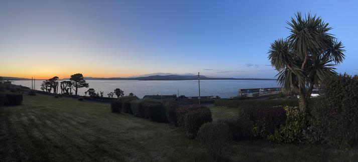 Panorama looking north across Dungarvan bay under a completely cloudless darkening blue sky. The sun has set to the west along the left hand edge of the picture turning the sky a golden colour on that side of the picture. 

The foreground is dark with some trees on the left in the middle distance silhouetted against the water of the bay, and Helvick harbour on the right partially obscured by a cordalyne australensis. 