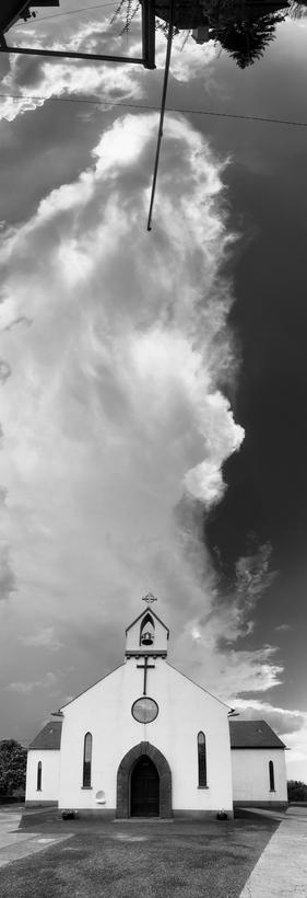 Vertical black white panorama photo rising up from a church, overhead clouds and with the barrier and trees  behind us topping off the image. 
