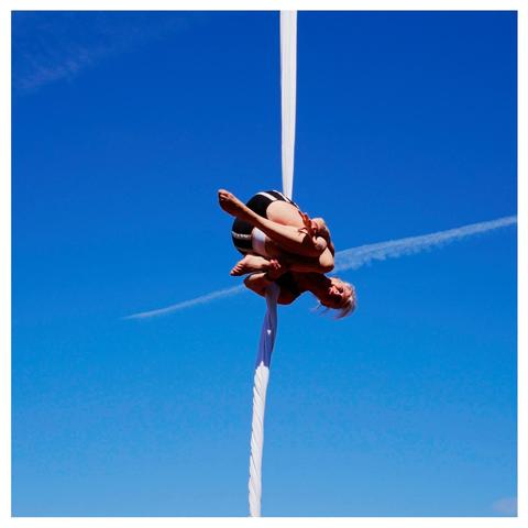 Suspended Tumble Circus performer in a ball knotted with the fabric against blue skies with one con trail passing behind her. 