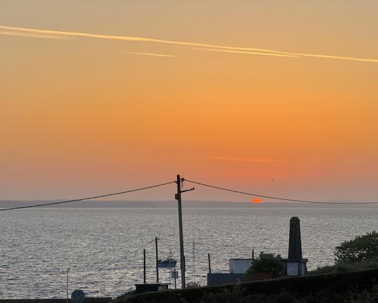 Sunrise at Helvick looking north east just as the sun breaks over the distant cliffs of the Copper Coast into a deep orange dawn sky. 

The Erin’s Hope monument is visible in the foreground. 