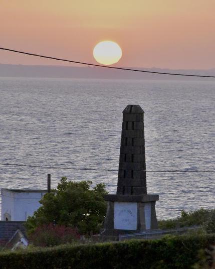 Sunrise at Helvick looking north east just as the full solar disc sits just touching the distant cliffs of the Copper Coast into a rapidly brightening  orange dawn sky. 

The Erin’s Hope monument is visible in the foreground just to the right of the sun. 