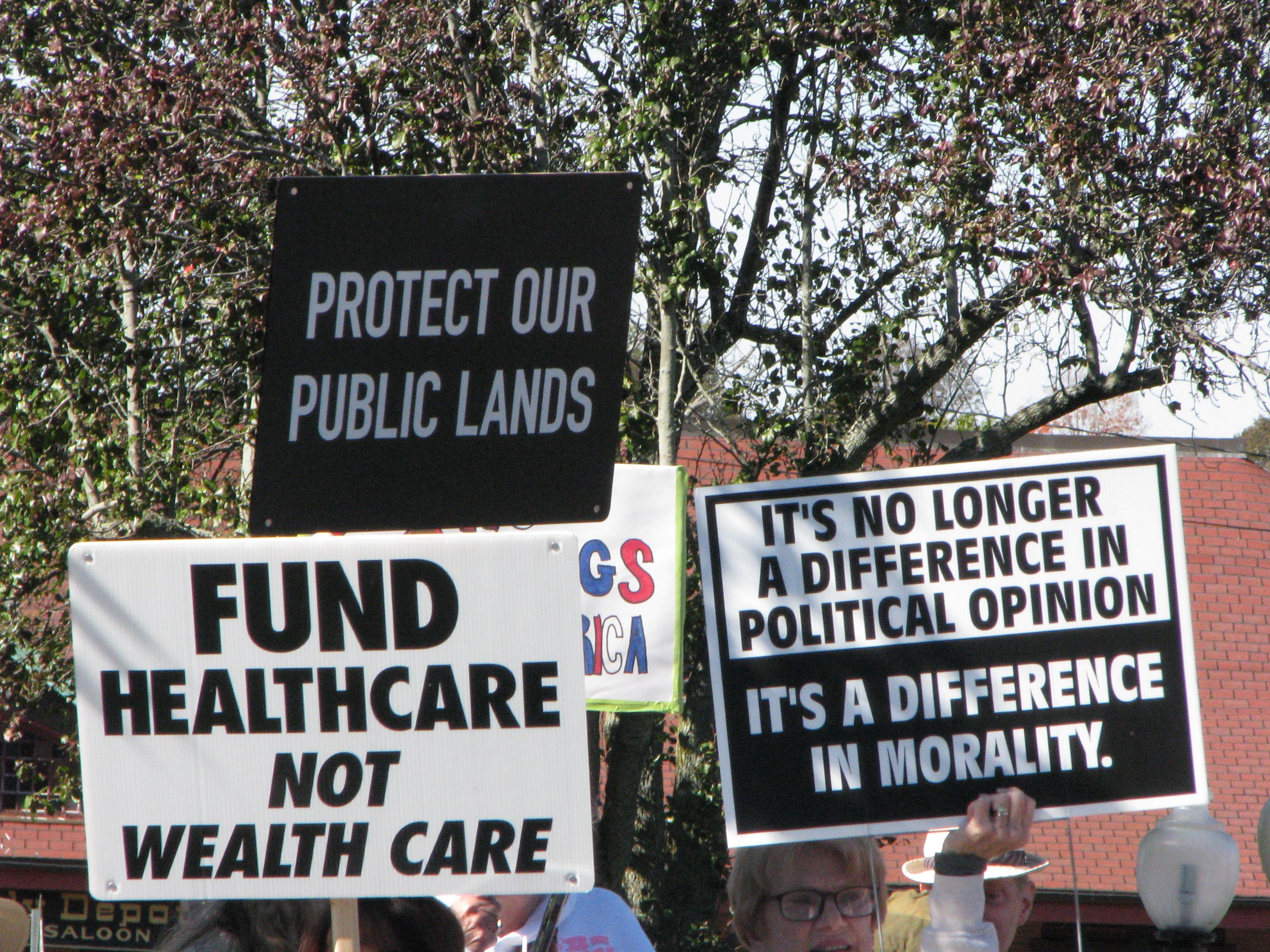 Multiple protest posters. One reads "protect our public lands". Another reads "Fund healthcare not wealthcare". A third reads "It's no longer a difference in political opinion, it's a difference in morality"