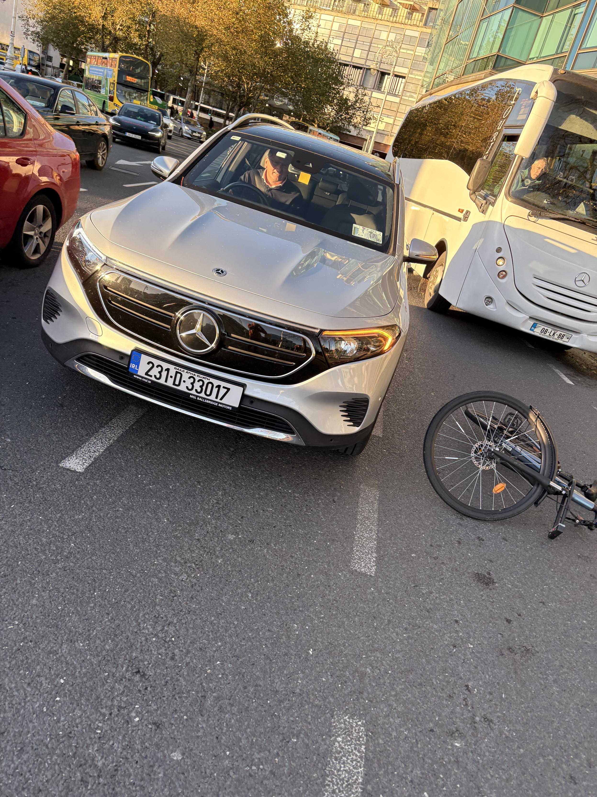 A silver Mercedes-Benz car parked on the street with a bicycle lying on its side nearby. Several other vehicles and a bus are in the background.
