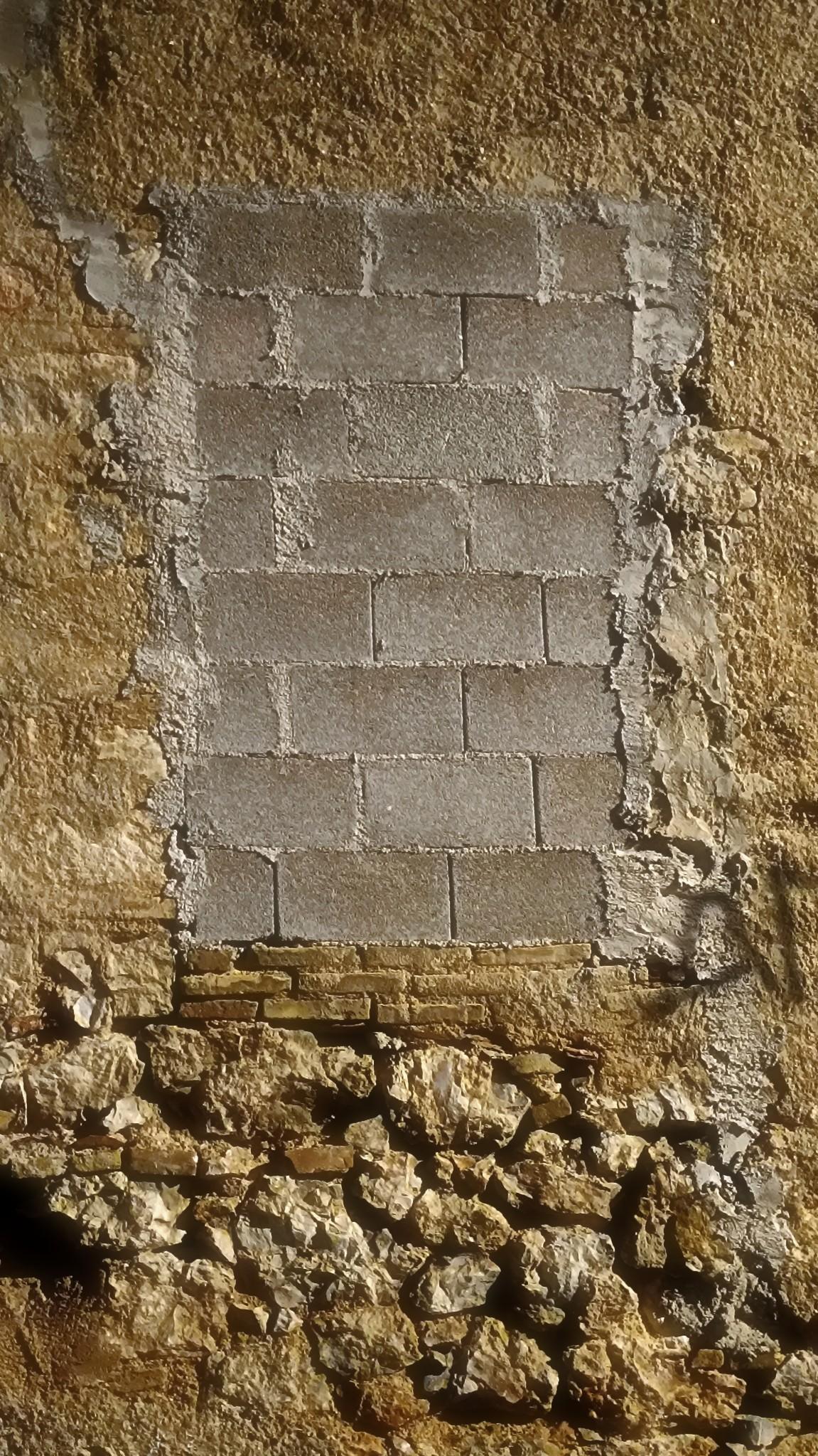 A window of an abandoned house, bricked with grey cement blocks, lit from the right and centered in the frame. A rectangular filled to the brim with rectangulars, in a rectangular. A few rows of thin clay bricks from another era are still in place where there once was a window sill, under the blocks. The wall around the window is roughly plastered. The lower part of it before the ground is naked, letting the old stonework see the light. There's a hole on the left, some stones are missing.
More events to the right (some writing in spray paint, but that's another photo).