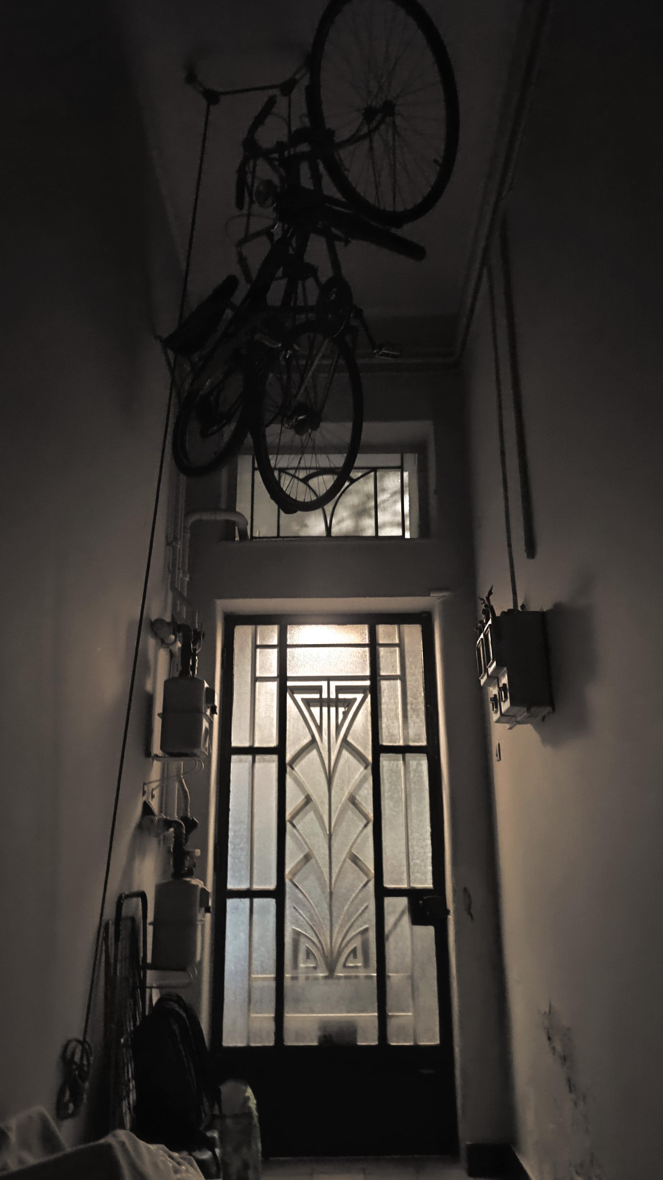 Photo of a narrow corridor ending at a door that leads outside, on the street. The light of a street lamp reaches inside, along with blue hints of dawn. Two bicycles are hanging above the door. A grocery carrier, a backpack and some more objects are left aside. Heat and electricity panels, new and old. 