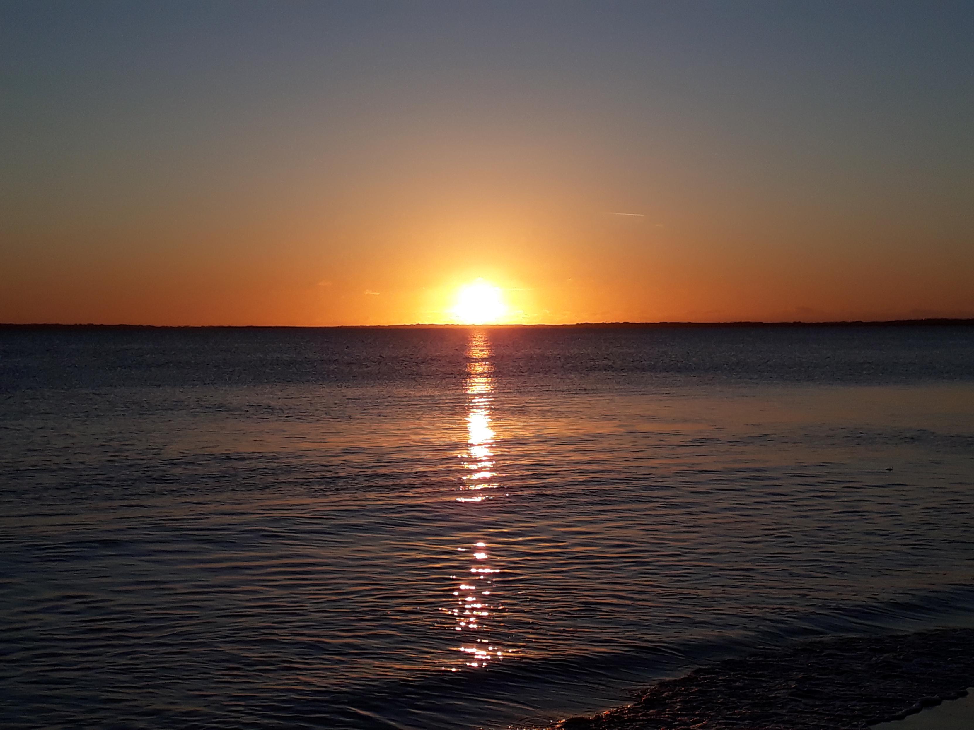 Photography of a sundown.
The sun is in the mid of the picture in the upper third.
the half of the sun is yellow-orange abd this colour spreads the whole width.
Above is a dark blue sky.
Below is dark curled water, which reflects the sun. on the roght bottom is a small bit beach