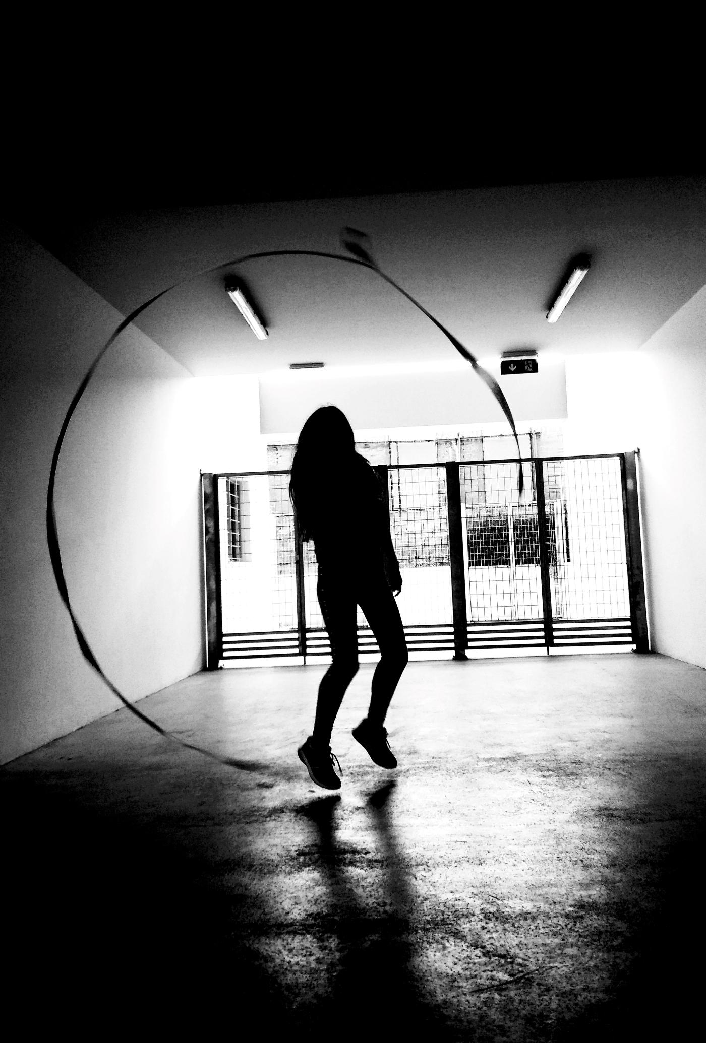 Black and white photo: The black figure of a young woman against the light behind her. Her feet are slightly over ground, as she draws a circle around her with a ribbon wand. She is standing in a shady, impersonal public space. Behind her there's an exit to the outer world, blocked by an iron gate.