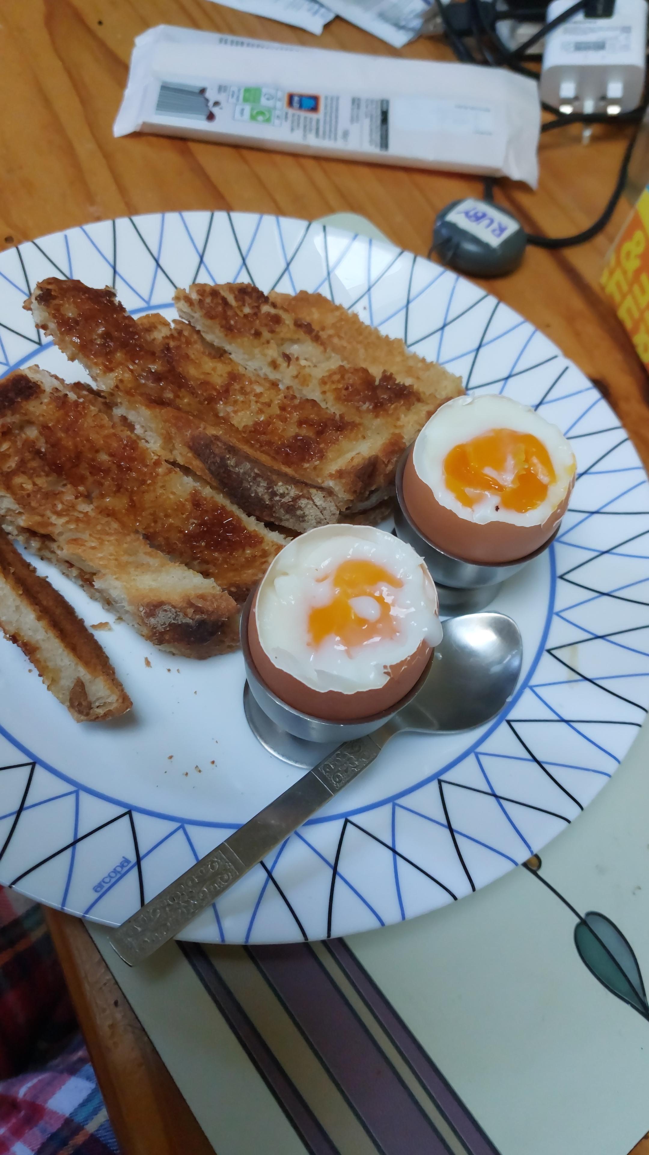 Two boiled eggs sit in silver eggcups on a plate, tops already removed, displaying the jammy soft yolks inside. There's a small pile of soldiers (buttered toast with marmite, cut into strips for dipping) and an old stainless steel teaspoon alongside.