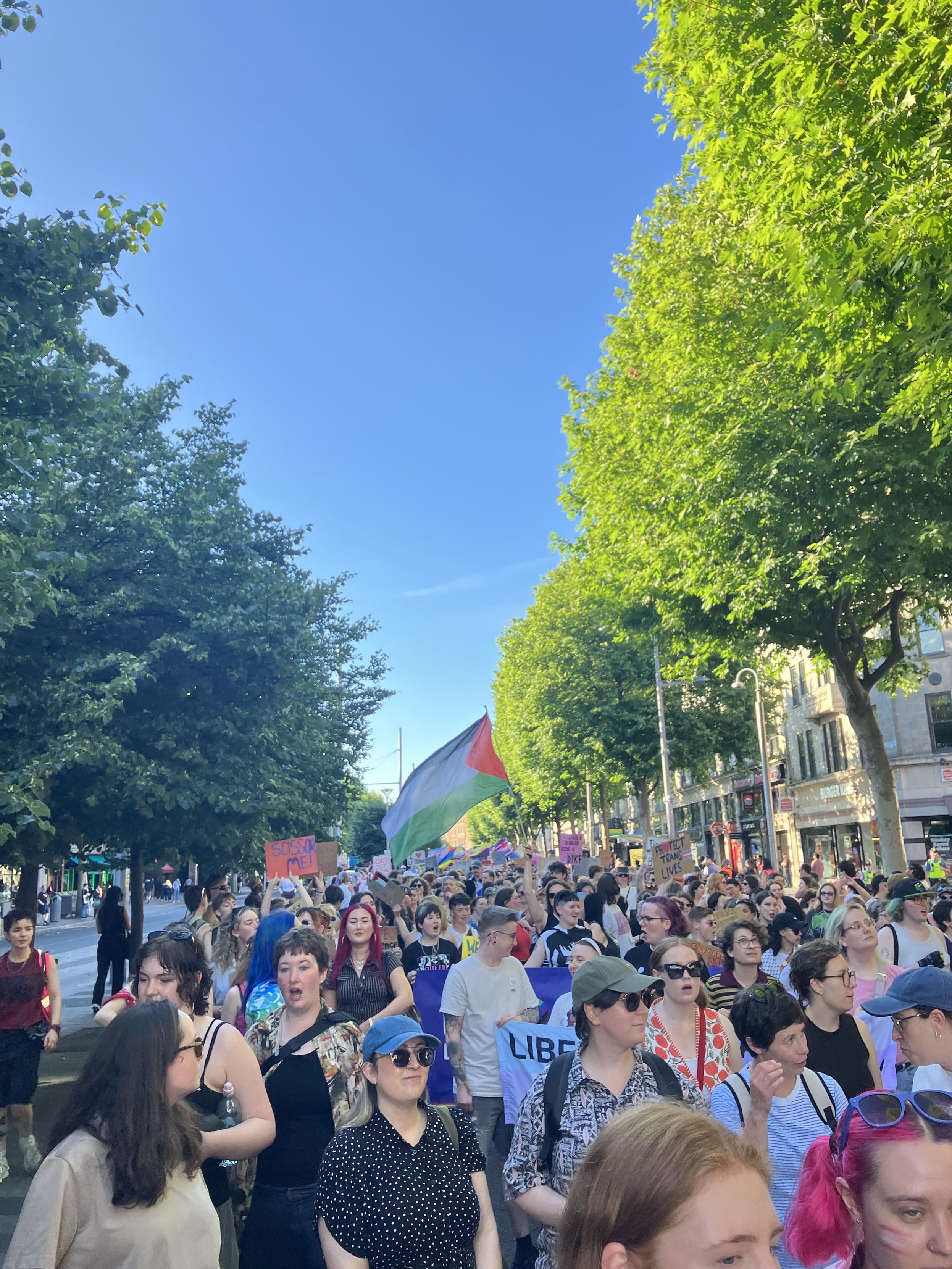 View of Dublin Dyke March 2026, taken on O'Connell Street, looking back from around half-way along the tree-lined street towards Parnell Street, with thousand of people, flags, and banners, under a beautiful clear blue sky.