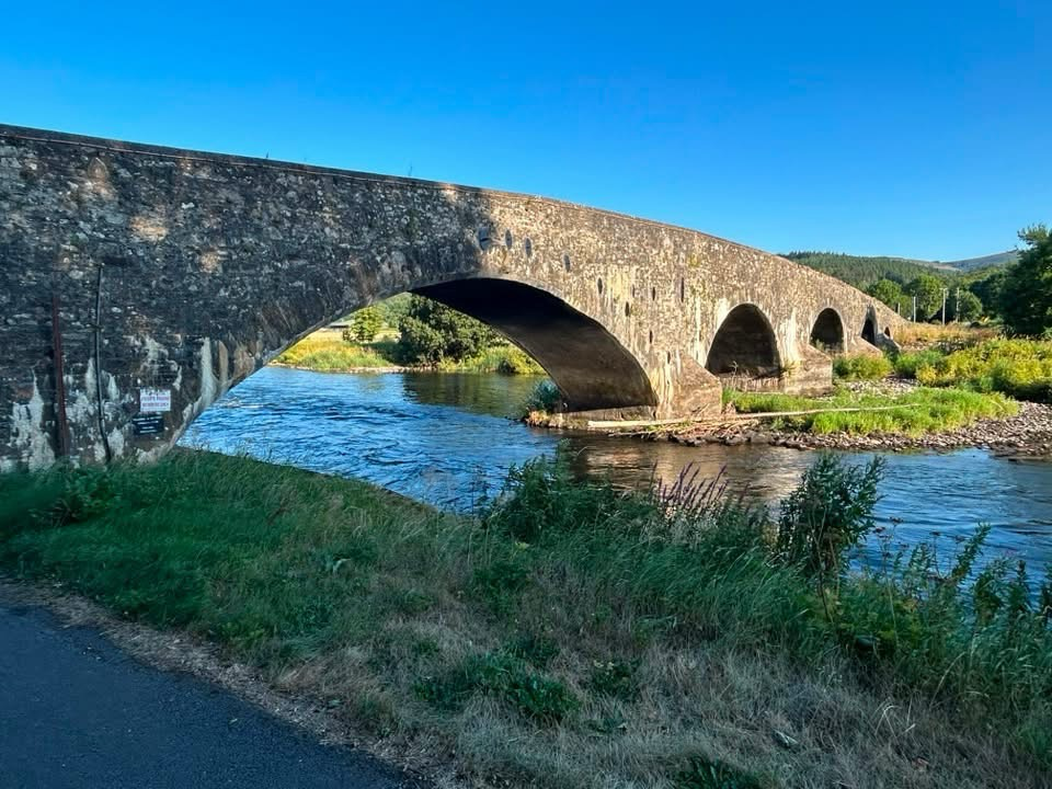 Colour photo of an old stone bridge with 4 arches crossing a shallow river. The sky is a vivid blue with no clouds. There is green grass on the banks of the river.