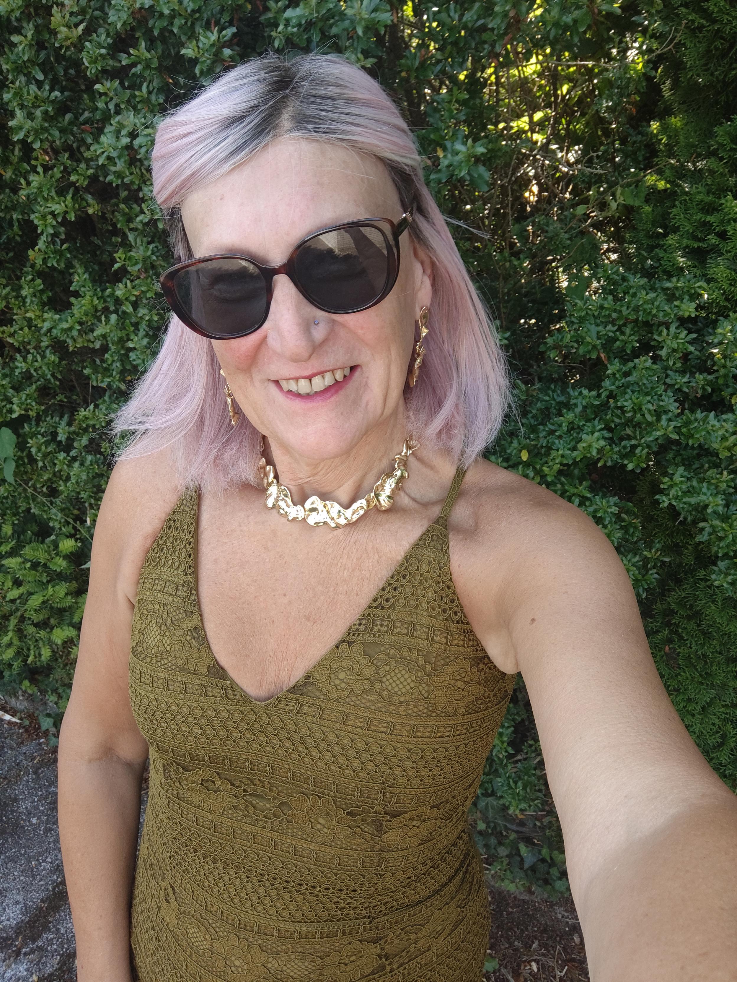 A pink haired woman standing in front of bushes wearing a green halter neck dress, chunky gold necklace, matching earrings and sunglasses.