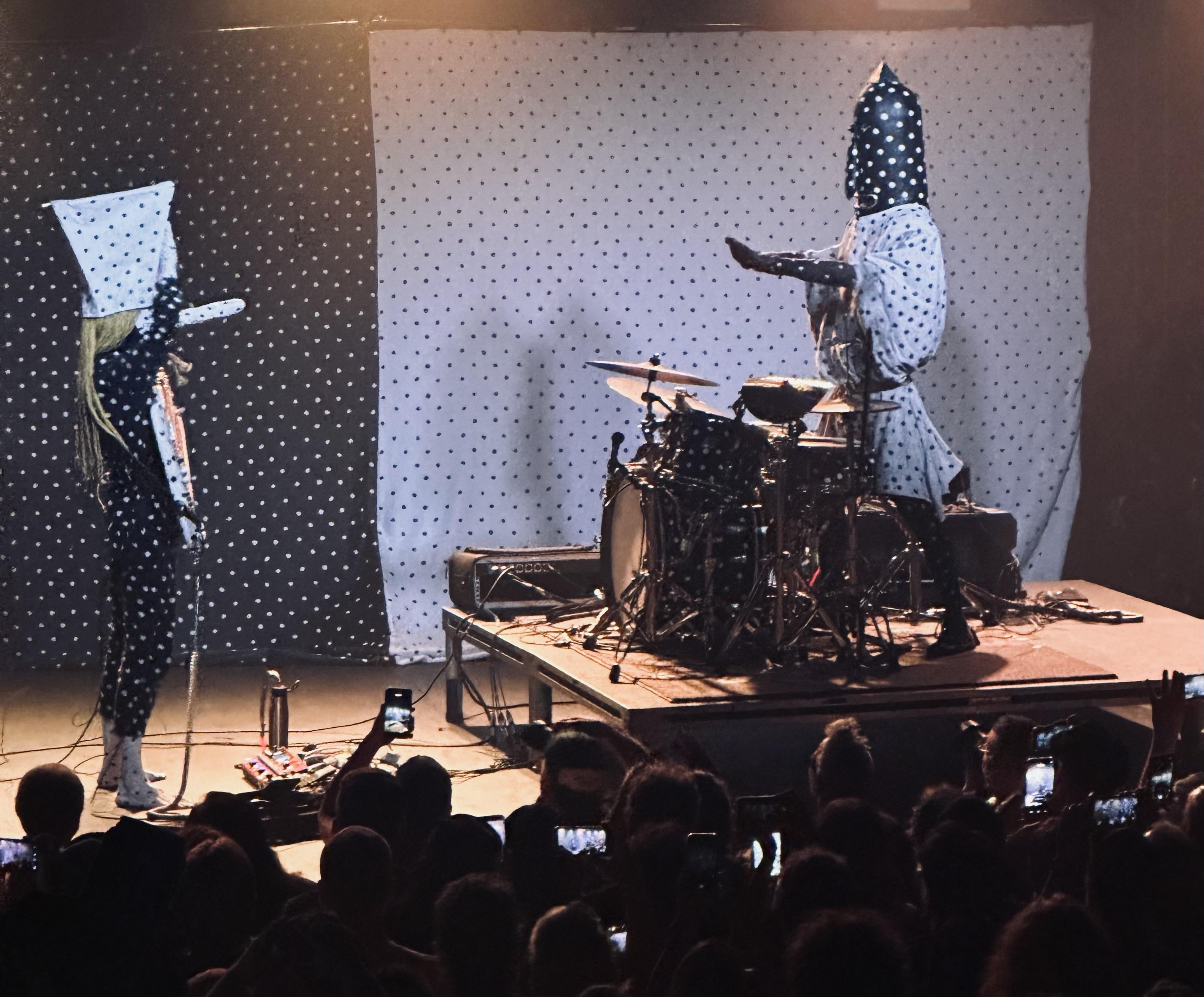 A live music performance featuring two musicians dressed in unique polka-dotted costumes, one playing a guitar and the other on drums. The background has a patterned fabric, and the audience is visible, holding up phones to capture the moment.